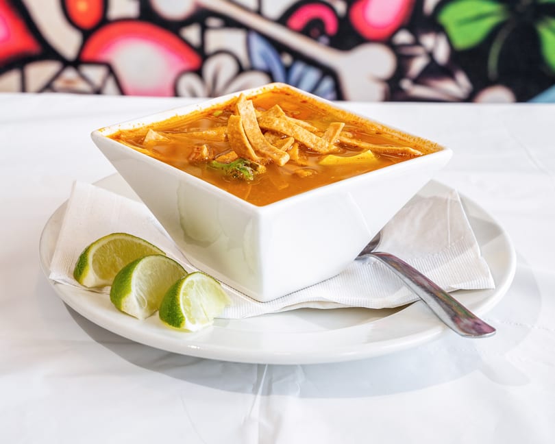 Tortilla soup in a white square bowl, garnished with tortilla strips and lime wedges.