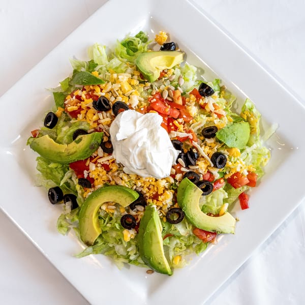 Fresh salad with lettuce, avocado, olives, tomatoes, cheese, sunflower seeds, and sour cream on a white plate.
