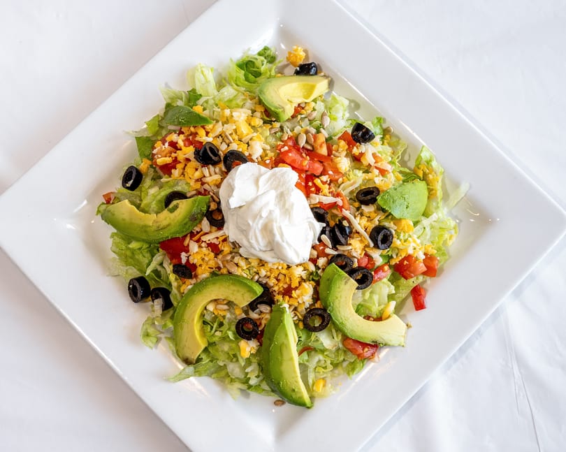 Fresh salad with lettuce, avocado, olives, tomatoes, cheese, sunflower seeds, and sour cream on a white plate.