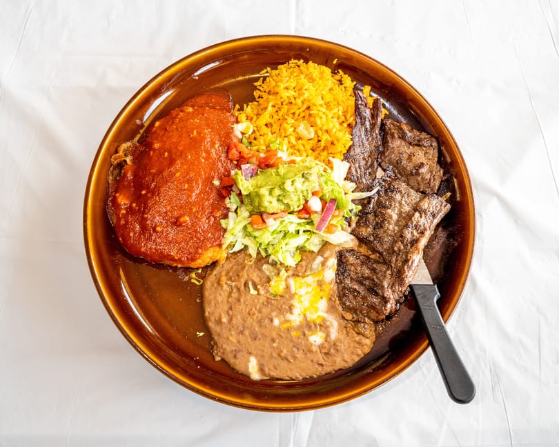 Large brown plate with grilled steak, yellow rice, refried beans, red sauce dish, and guacamole salad.