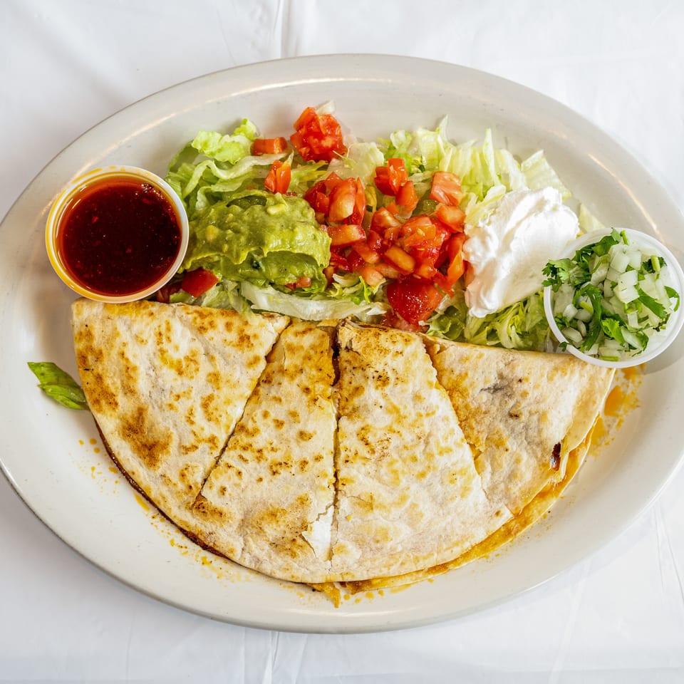 Quesadilla wedges with shredded lettuce, guacamole, diced tomatoes, sour cream, onions, and red sauce on a white plate.