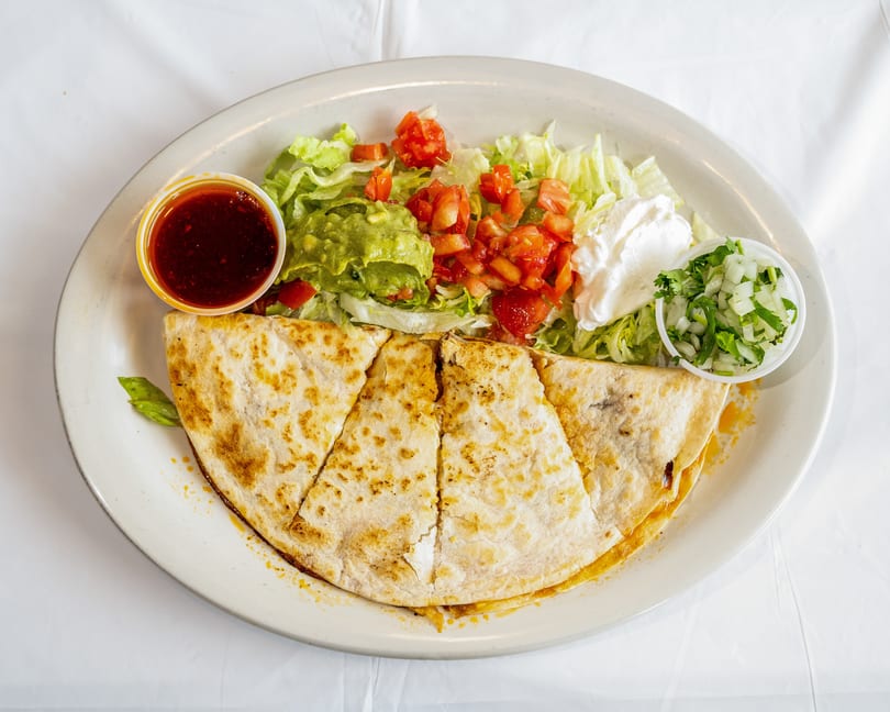 Quesadilla wedges with shredded lettuce, guacamole, diced tomatoes, sour cream, onions, and red sauce on a white plate.