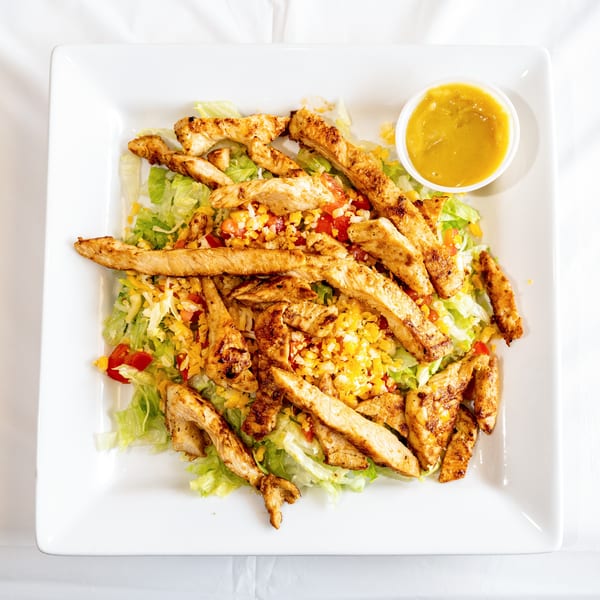 Grilled chicken salad with lettuce, cheese, tomatoes, corn, and a side of dressing on a white plate.