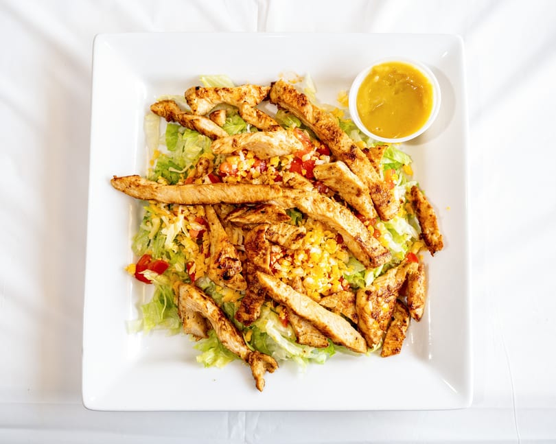 Grilled chicken salad with lettuce, cheese, tomatoes, corn, and a side of dressing on a white plate.