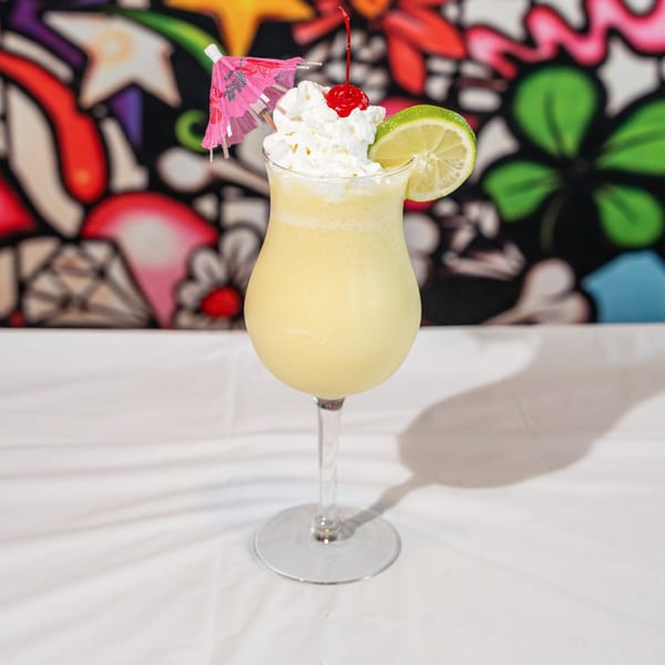 Tall glass of yellow frozen drink with whipped cream, cherry, lime, and pink umbrella.