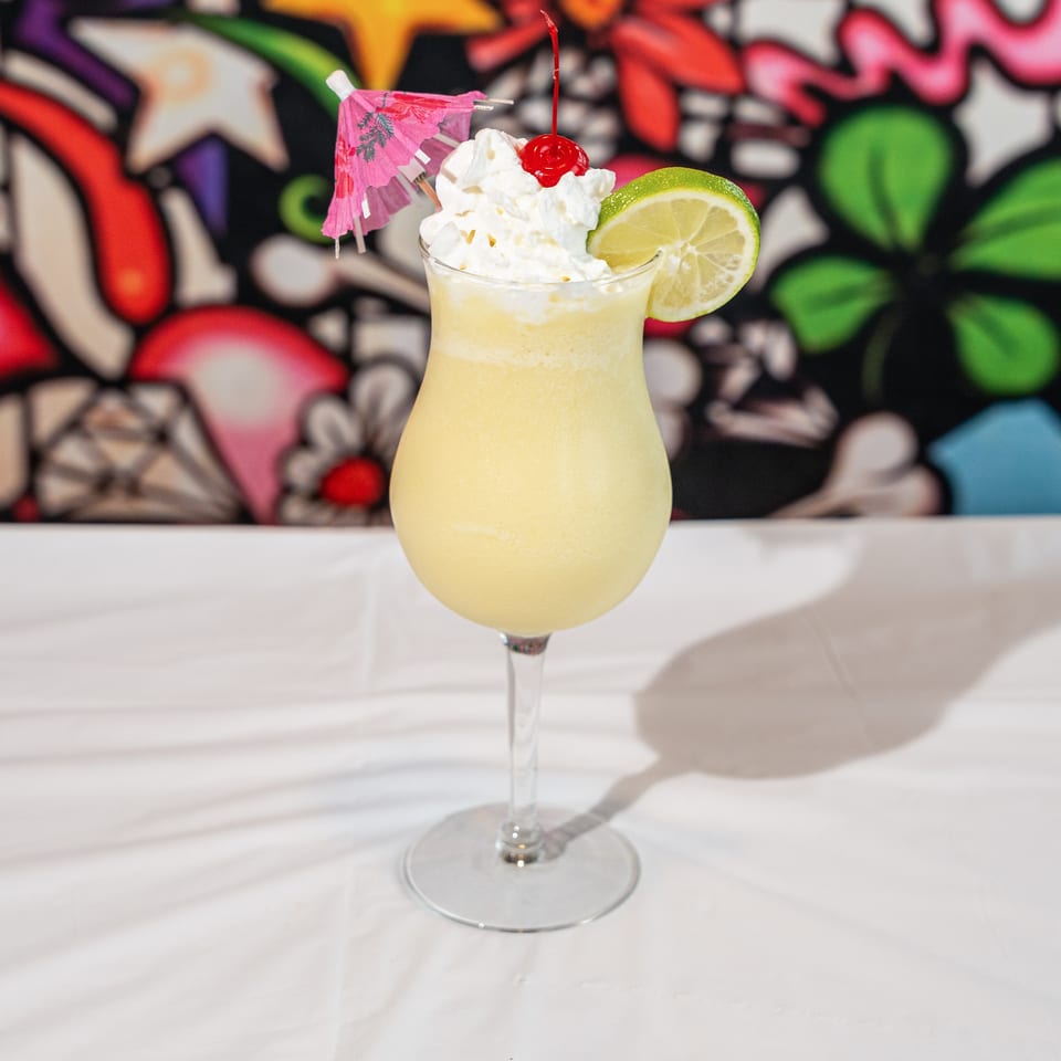Tall glass of yellow frozen drink with whipped cream, cherry, lime, and pink umbrella.