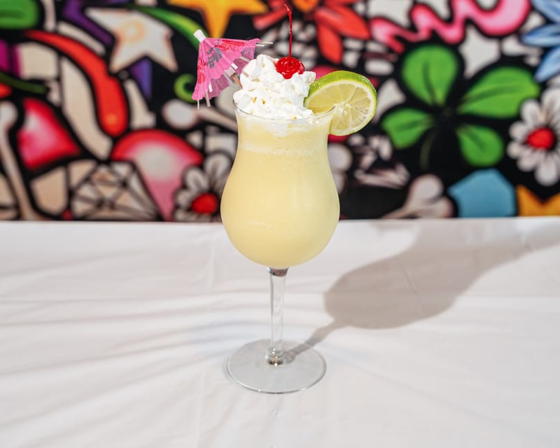 Tall glass of yellow frozen drink with whipped cream, cherry, lime, and pink umbrella.