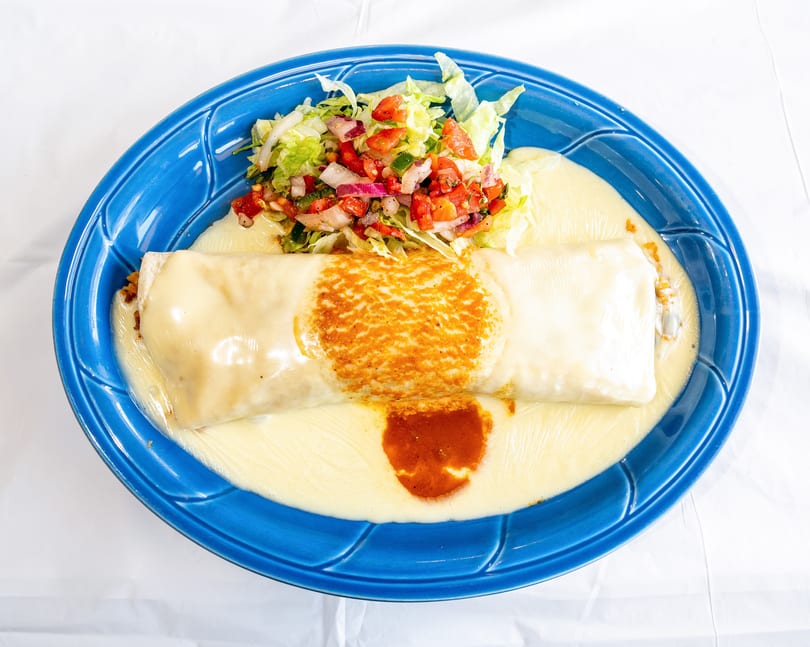 Large burrito smothered in white cheese sauce, with pico de gallo and lettuce on a blue plate.